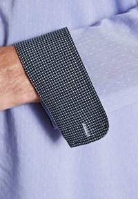 Helles blaues Hemd mit kleinen, strukturierten Punkte. Der Ärmelabschluss besteht aus einem kontrastierenden dunkelblauen Stoff mit weißen Kreismustern.