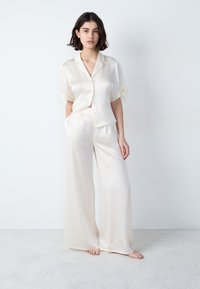 Zijden pyjama-set in off-white met een kort-gevoerde button-up top en wijde broekspijpen, zachte textuur en subtiele ruches.