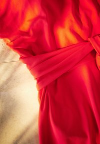 Tessuto rosso drappeggiato in morbide pieghe con dettaglio di cintura attorcigliata, che mostra una texture liscia. Gradiente caldo che sfuma all'arancione ai bordi.