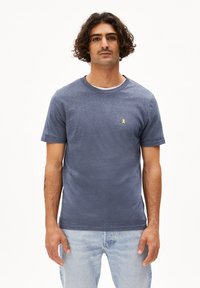 T-shirt blu navy con scollo rotondo in tessuto testurizzato, caratterizzato da un piccolo logo a nastro giallo sul petto. Abbinato a jeans azzurri.