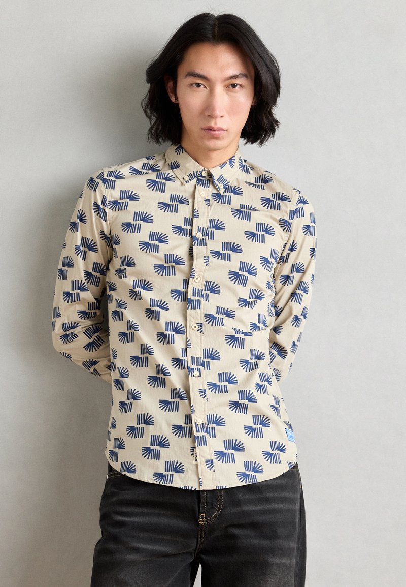 Beige skjorte med knapper og et blåt geometrisk bladmønster, lange ærmer og en buet kant. Sammen med mørke jeans.