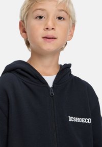 Blondé dieťa oblečené v čiernej mikine na zips s logom "DCSHOECO", pod ňou biela košeľa, na prázdnom svetlom pozadí.