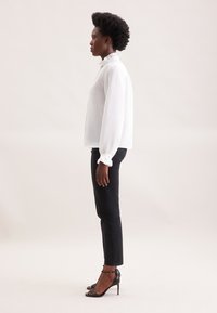 Weißes Blusenhemd mit plissiertem Kragen, langen Ärmeln und Manschettendetails, kombiniert mit schwarzen Slim-Fit-Jeans und schwarzen Riemchenheels.