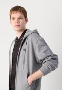 Grijze zip-up hoodie van zacht materiaal, met een voorzak, trekkoordcapuchon en geborduurd logo op de linkerborst.