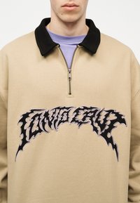 Κοντινό πλάνο ενός ατόμου που φοράει μπεζ πουλόβερ με μαύρο γιακά και εμπρόσθιο φερμουάρ, με έντονο μαύρο γραφικό κείμενο στο στήθος.