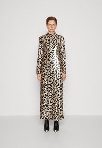 Maxi mekko leopardikuvioinnilla, jossa on pitkät hihat, korkea kaula-aukko ja kiiltävä paljettil fabric. Malli käyttää mustia nilkkureita, joissa on valkoisia yksityiskohtia.