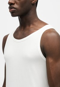 Nahaufnahme eines Mannes, der ein schlichtes weißes ärmelloses Tanktop vor einem schlichten hellen Hintergrund trägt.