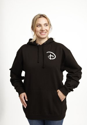 Donna con felpa nera con cappuccio e logo bianco Disney "D", mani in tasca, sorridente e che guarda di lato su sfondo bianco semplice.