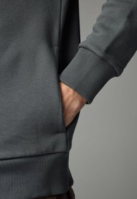 Sweat-shirt gris foncé avec poignets et ourlet côtelés. Une main est placée dans une poche latérale, mettant en valeur le design décontracté et la texture du vêtement.