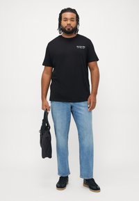 Man met dreadlocks, gekleed in een zwart graphic t-shirt, lichtblauwe jeans, zwarte sneakers, en draagt een zwarte tas tegen een effen witte achtergrond.