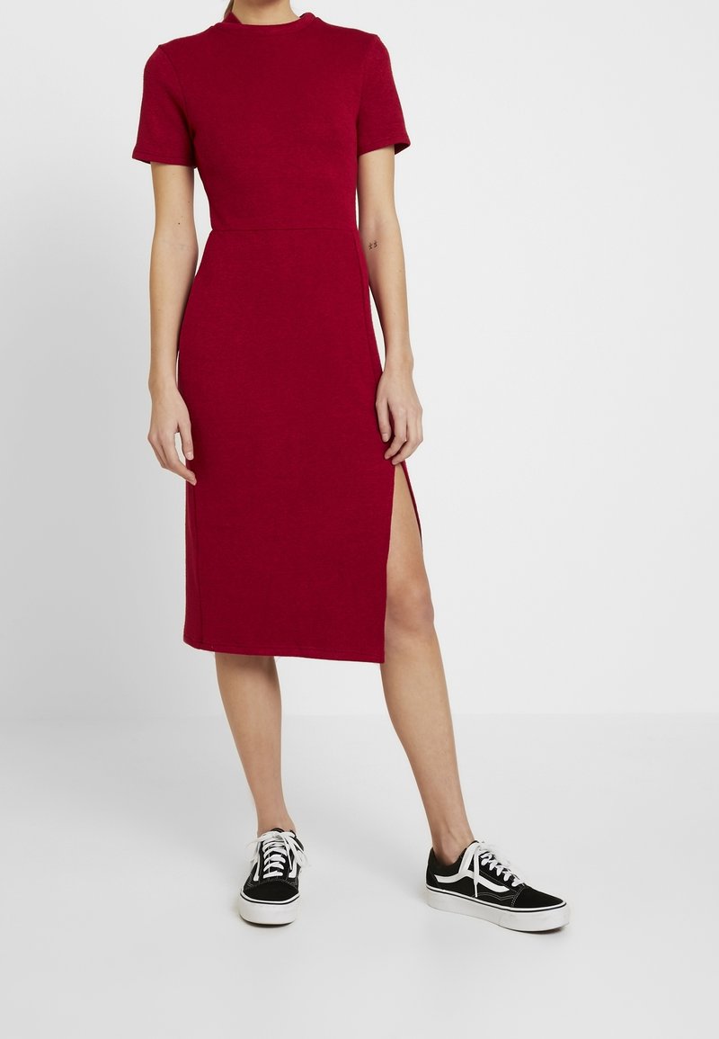 Femme portant une robe rouge mi-longue à manches courtes avec une fente latérale, associée à des baskets noires et blanches, debout sur un sol blanc.