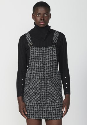 Suéter negro de cuello alto combinado con un vestido de overoles a cuadros que cuenta con dos bolsillos frontales y clips metálicos ajustables.