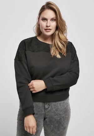 Black sweatshirt featuring textured black lace accents on the shoulders, ribbed cuffs, and a relaxed fit. Paired with gray jeans.