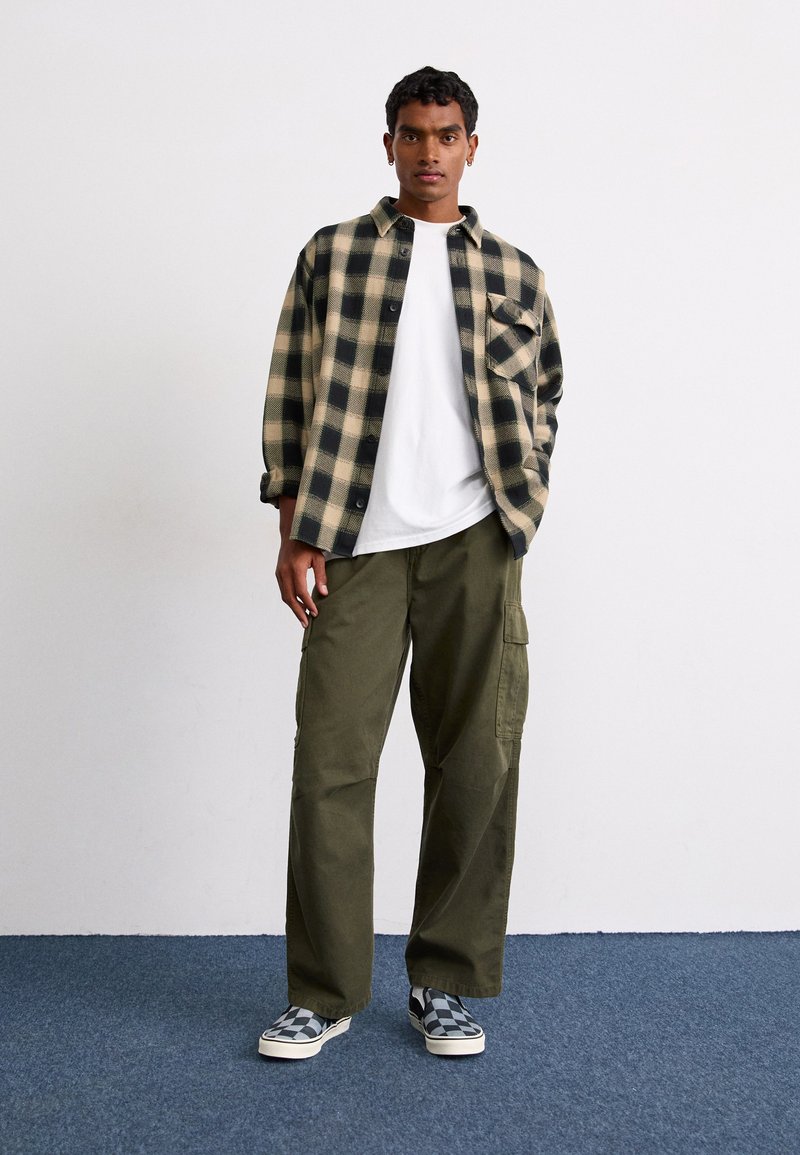 Camisa de franela a cuadros en negro y marrón, camiseta blanca debajo, pantalones cargo en verde oliva, zapatillas de slip-on a cuadros en blanco y negro.