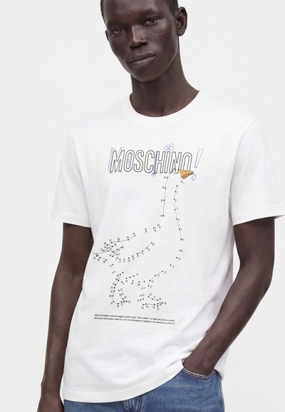 Άνδρας που φοράει λευκό t-shirt Moschino με γραφικό με κουκκίδες που συνδέουν τις τελείες σε παπάκι και μπλε τζιν, με απλό φόντο.