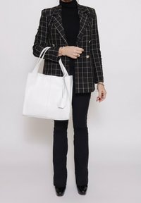 Borsa a spalla in pelle bianca con forma piatta, caratterizzata da cuciture laterali e un semplice dettaglio tag. Indossata con un blazer a quadri neri e pantaloni aderenti.