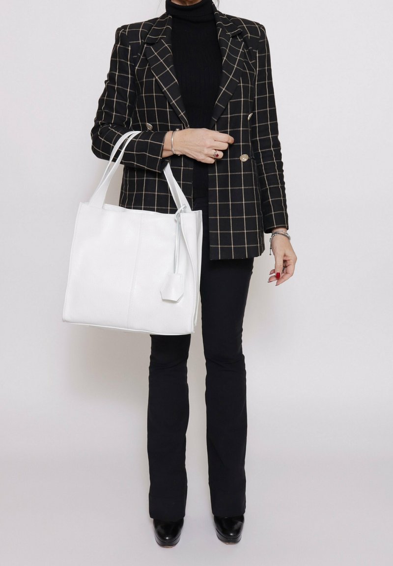 Borsa a spalla in pelle bianca con forma piatta, caratterizzata da cuciture laterali e un semplice dettaglio tag. Indossata con un blazer a quadri neri e pantaloni aderenti.