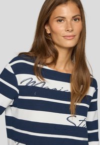 Junge Frau mit langen braunen Haaren, die ein marineblau-weiß gestreiftes Oberteil mit dekorativem Text und Perlenakzenten trägt.