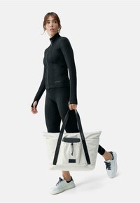 Balta tote soma ar melniem rokturiem, ar savelkam ām aizdares un priekšējo kabatu. Modelis valkā melnu zip-up jaku un melnas legingu bikses.