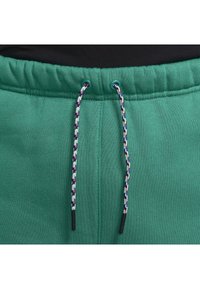 Tealgröna sweatpants med resårmidja och flerfärgade flätade dragsnören hängande framme mot en svart bakgrund.