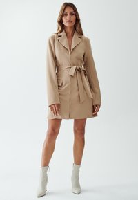 Beige takki mekko, jossa on vyö, kauluskrae, pitkät hihat ja kaksi etutaskua. Yhdistetty vaaleanharmaisiin nilkkureihin yksinkertaista taustaa vasten.