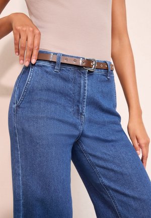 Nő kék, magas derekú farmernadrágot visel oldalzsebekkel és barna övvel, bézs ujjatlan felsővel kombinálva.