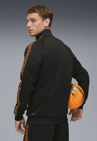 Schwarze Sportjacke mit orangefarbenen Akzenten, hohem Kragen und elastischen Bündchen. Das Modell hält einen orangefarbenen Fußball an seiner Seite.