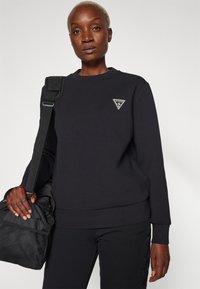 Felpa nera in misto cotone, con scollatura rotonda e una toppa con logo triangolare. Indossata con pantaloni neri abbinati e una borsa.