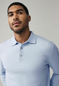 Himmelblaues Strickpolohemd mit Kragen, zwei Knöpfen und langen Ärmeln. Glatte Textur, figurnaher Schnitt, minimaler Akzent.
