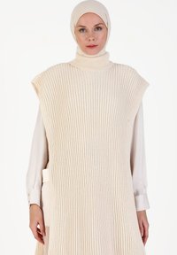 Tunique en maille côtelée beige avec un col montant, silhouette oversize et manches courtes. Portée par-dessus un haut blanc à manches longues avec des accents de boutons.