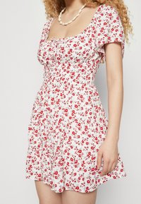 Vit blommig klänning med röda blommor, korta puffärmar, fyrkantig halsringning och en åtsittande överdel med rynkad textur vid midjan.