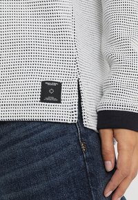 Sudadera blanca con un patrón, acabado texturizado, puños negros y etiqueta lateral. Llevada sobre unos vaqueros azul oscuro, mostrando un estilo casual.