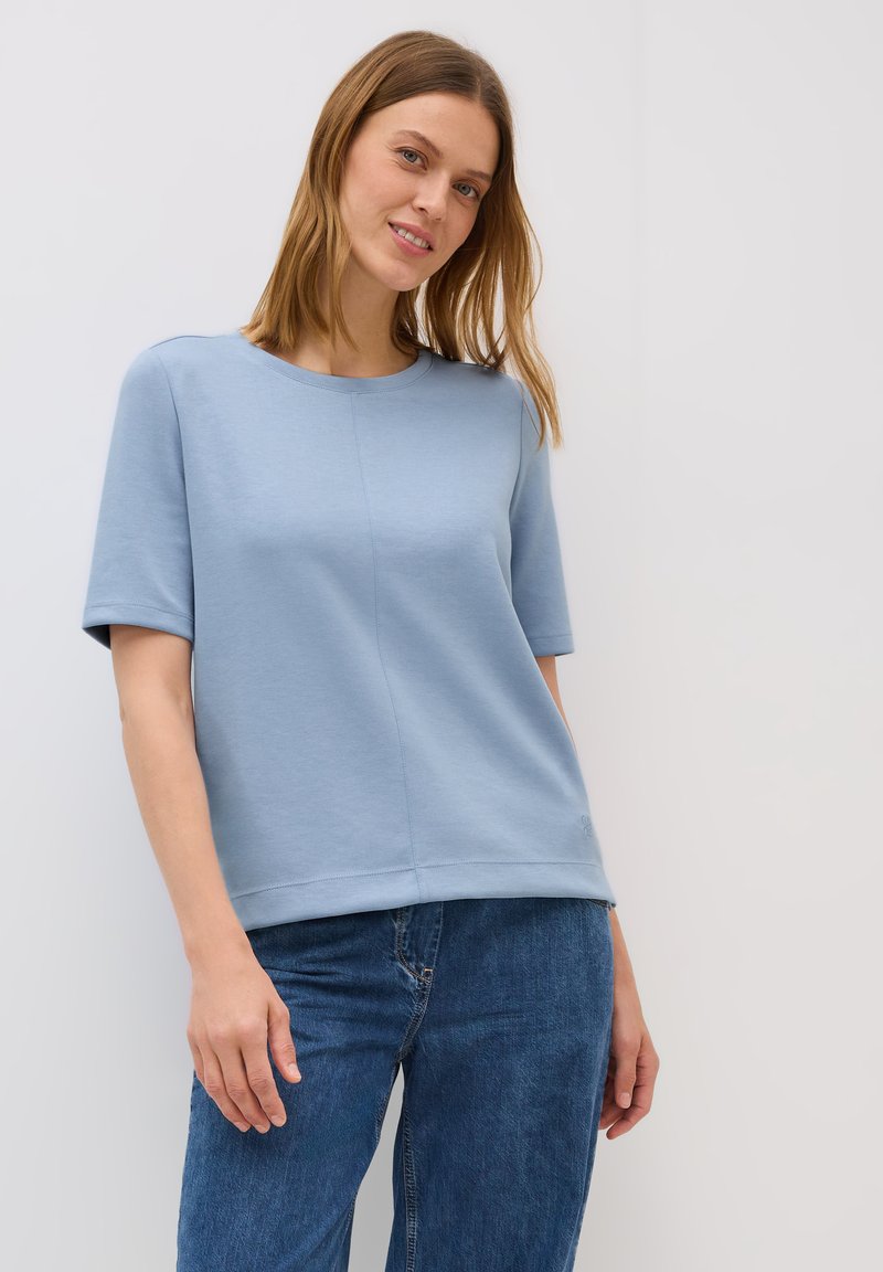 Model poseert in een lichtblauwe korte mouwen top, gecombineerd met blauwe jeans, glimlachend tegen een effen witte achtergrond.