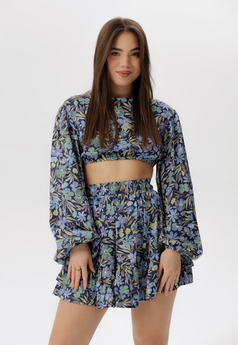 Conjunto de top corto floral y falda de volantes, con patrones florales en azul y verde sobre un fondo oscuro, con mangas largas y cintura elástica.