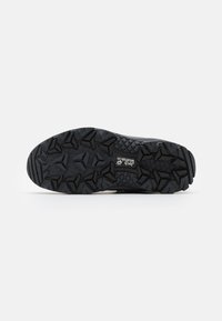 Semelle de chaussure de randonnée noire et robuste avec des crampons triangulaires profonds et le logo "Jack Wolfskin" au centre sur un fond blanc uni.