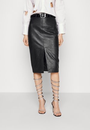 Femme portant une jupe crayon noire en similicuir à texture crocodile avec une fente à l'avant, une chemise blanche à manches longues avec des découpes en forme de cœur, et des sandales à talons hauts à brides.