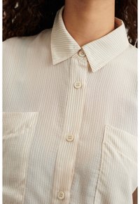 Camicetta color beige chiaro con strisce verticali sottili, colletto e una tasca sul petto. Presenta quattro bottoni e un tessuto testurizzato.