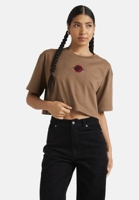 T-shirt marron court en coton, avec un motif d'œil brodé rouge et or. Porté avec un pantalon en velours côtelé noir taille haute.