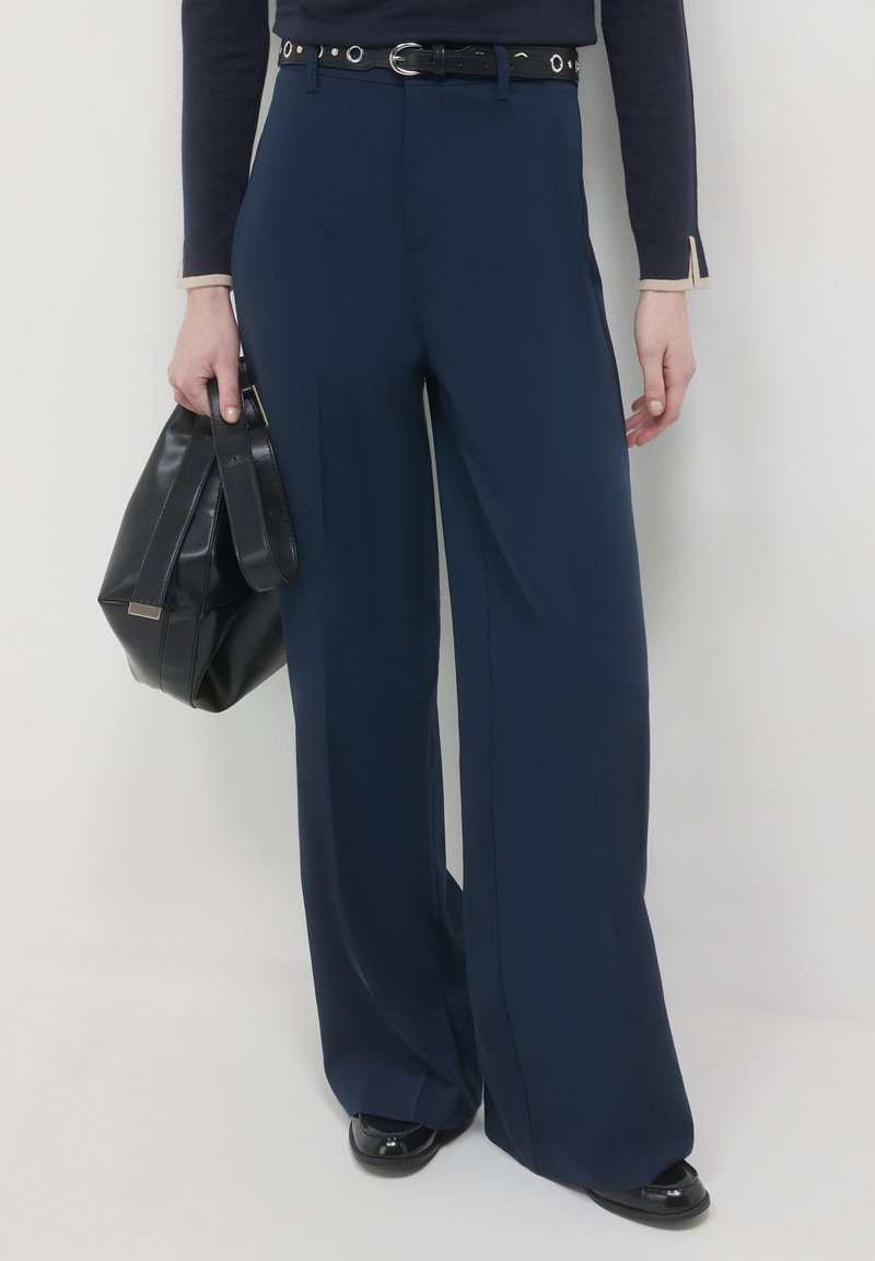 Navy hochtaillierte weit geschnittene Hose aus glattem Stoff, ausgestattet mit einem schwarzen Gürtel mit silberner Hardware, kombiniert mit einer schwarzen Handtasche.