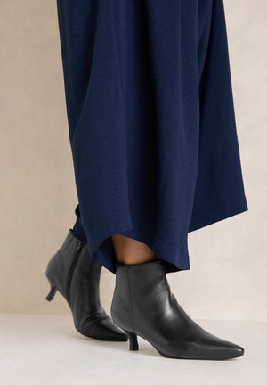 Personne portant des bottines en cuir noir à talons bas et une longue jupe texturée bleu foncé.