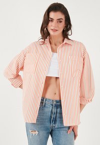 Pruhovaná košeľa s oranžovými a bielymi pásmi, s dvoma náprsnými vreckami, s rolovanými rukávmi a vo voľnom strihu, nosená cez biely crop top.