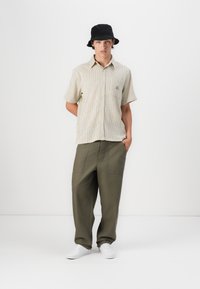Stripete kortermet skjorte i beige med lomme detaljer, parret med løse olivengrønne bukser og sort bøttehatt. Hvite sneakers fullfører looken.