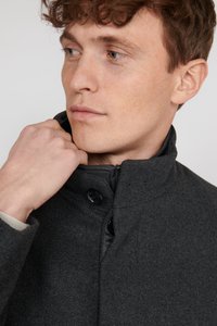 Dunkelgrauer Wollmischmantel mit hohem Kragen, Reißverschluss und schwarzen Knöpfen. Glatte Textur mit strukturiertem Design und minimalistischem Stil.