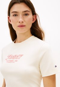 Κοντομάνικη κρεμ μπλούζα με κόκκινο λογότυπο "TOMMY HILFIGER, CO.". Διαθέτει στρογγυλή λαιμόκοψη και μικρή λεπτομέρεια σημαίας στο μανίκι. Λείο ύφασμα.