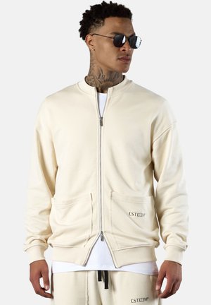 Man wearing cream-colored zip-up jacket and matching pants, with sunglasses and visible neck tattoos, standing against a plain light background.