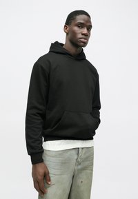 Μαύρη φούτερ με κουκούλα και ευρύχωρη μπροστινή τσέπη, με ριμπ μανίκια και στρίφωμα, φορεμένη πάνω από λευκό πουκάμισο. Το μοντέλο ποζάρει έναντι ενός απλού φόντου.