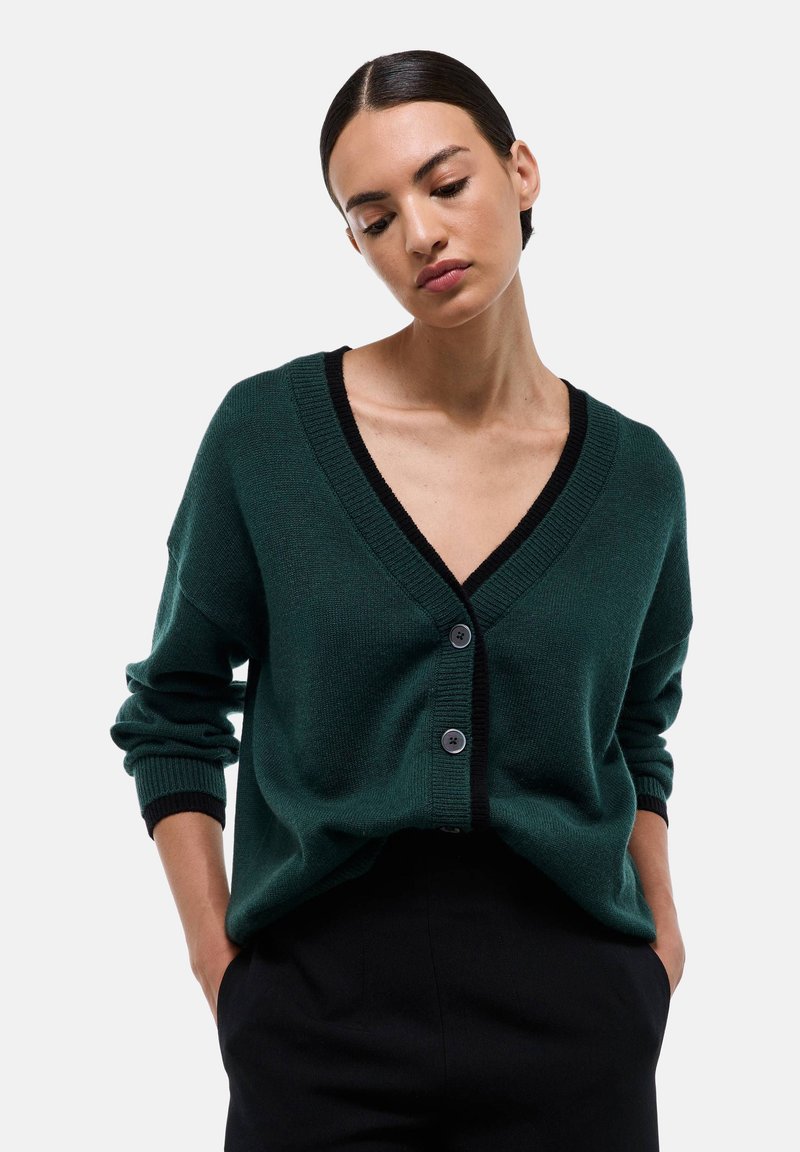 Gilet en tricot vert avec un décolleté en V, des bordures noires et trois boutons. Présente des épaules tombantes et des détails de poignets côtelés. Design simple et court.