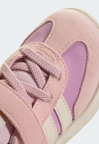 Detailní záběr na růžovou a fialovou tenisku s texturovaným materiálem, sametovými překryvy, křížově zavázanými tkaničkami a suchým zipem na světlém pozadí.