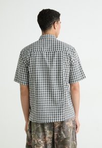 Cămașă cu mâneci scurte în pattern cu carouri alb-negru, din material ușor, cu gât button-down, purtată cu pantaloni scurți cu model de camuflaj.