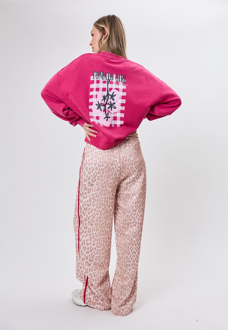 Kvinde i pink sweatshirt med teksten "Tomato Girl" og grafik, kombineret med beige leopardprint vidde bukser og hvide sneakers, stående med hænderne på hoften.